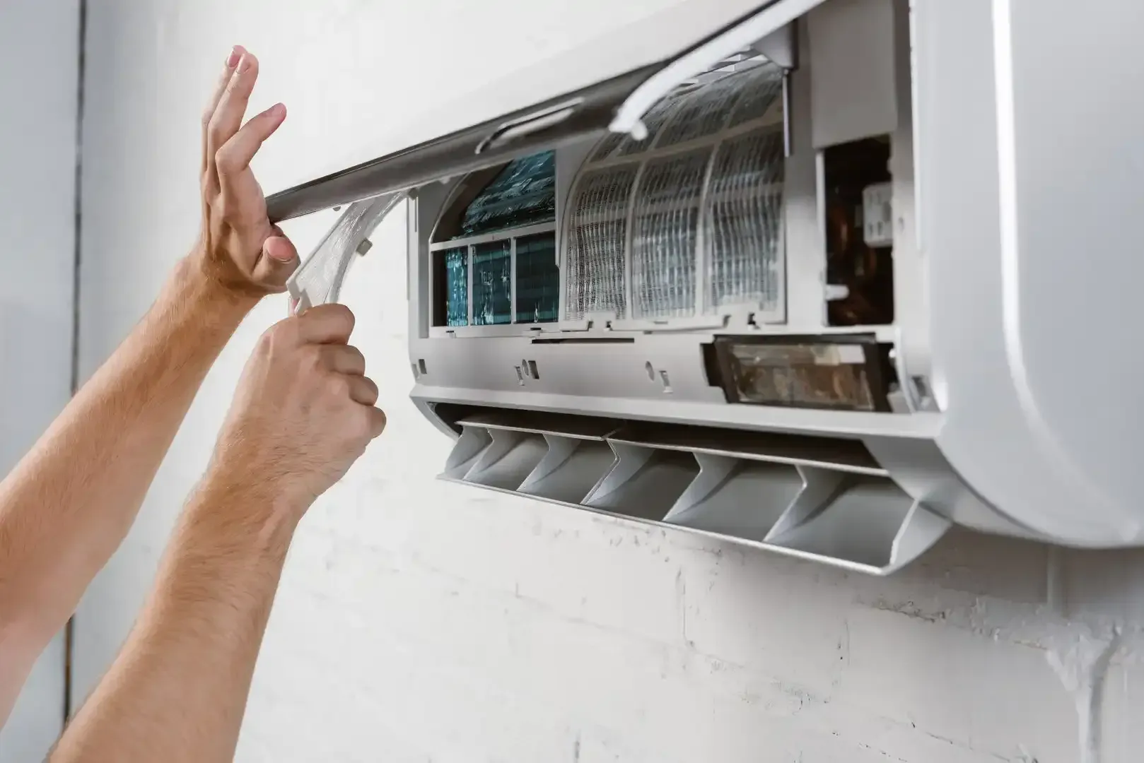 Hands cleaning an air conditioner unit, removing filter, white wall background.