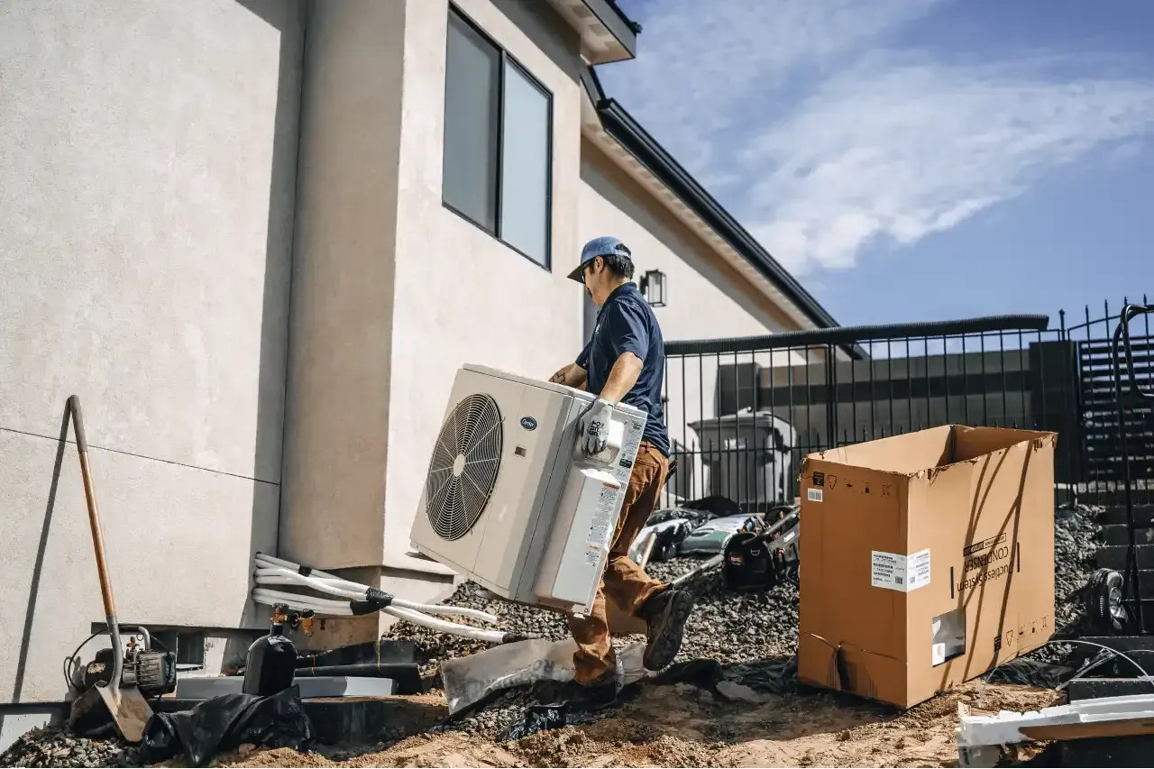 Man carrying a new air conditioning unit beside a house, under a blue sky. Cardboard box and debris nearby.