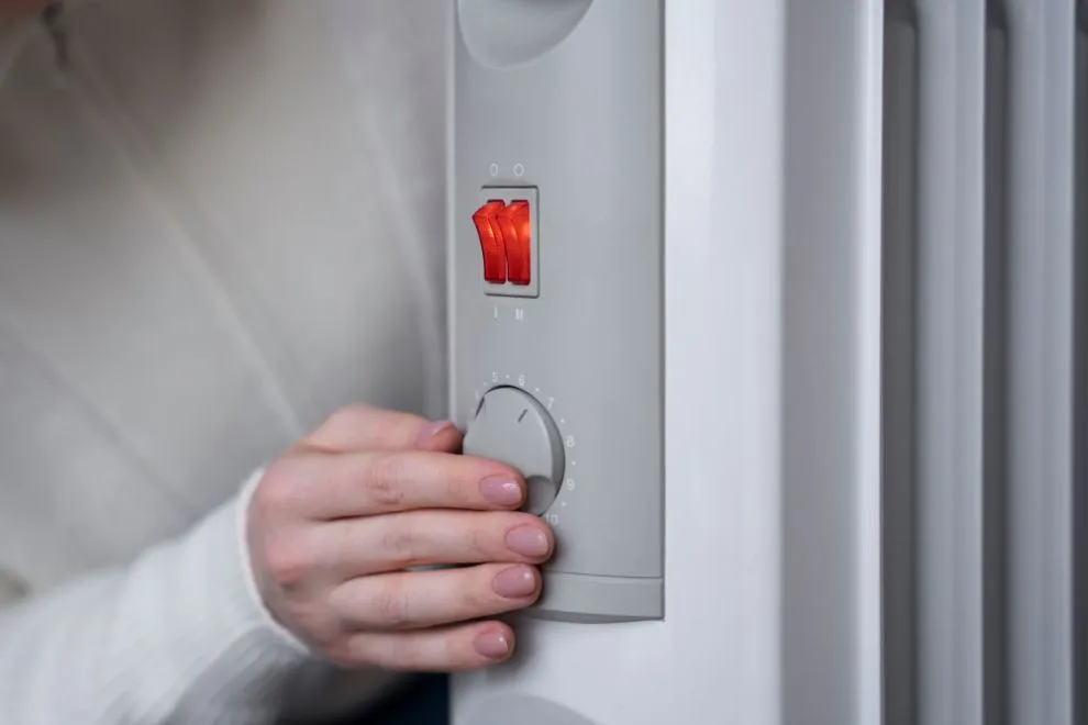Person adjusting the temperature dial on a white oil-filled radiator; red power switch is on.