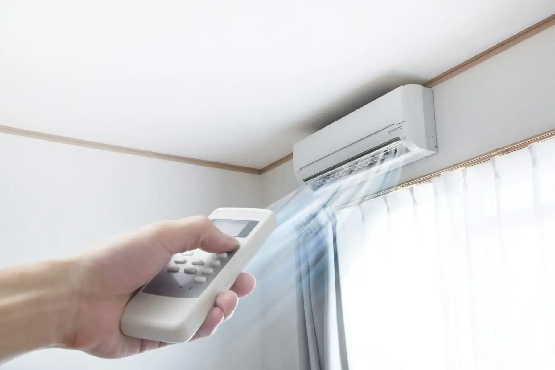 Hand holding a remote control pointing at a white air conditioner on a wall, with cool air blowing out.