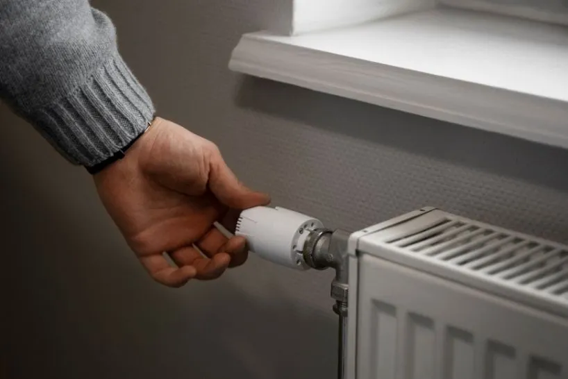 Hand turning a white thermostat dial on a radiator near a window.